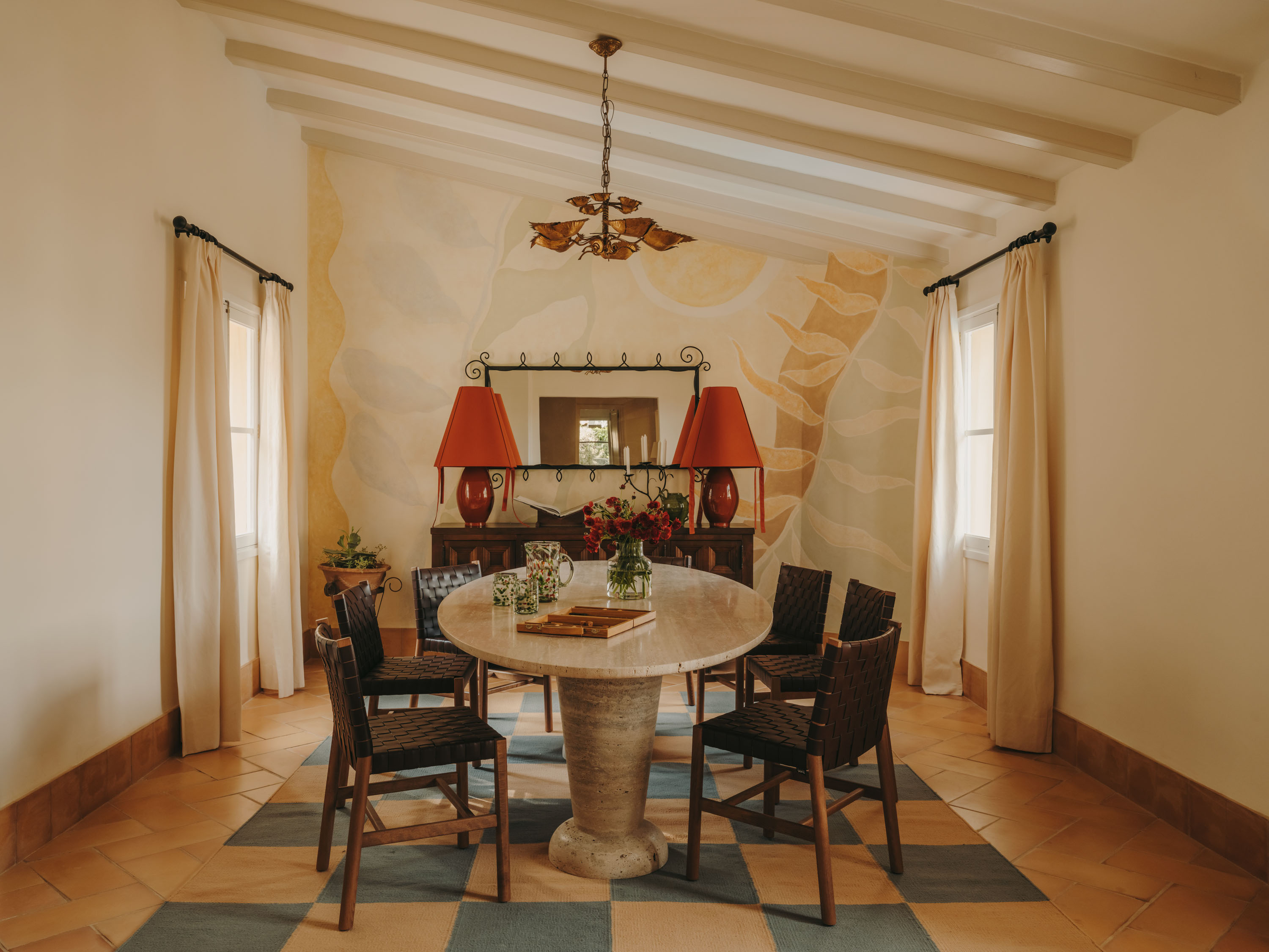 A warmly decorated apartment with heritage bones, checkered terracotta floors covered in a checkered blue and cream rug, marble table, red table lamps, and a cream-washed interior.