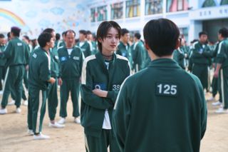 A woman (Won Ji-an) talks to a man as they stand near a crowd of people in matching green numbered tracksuits, in 'Squid Game' season 2.