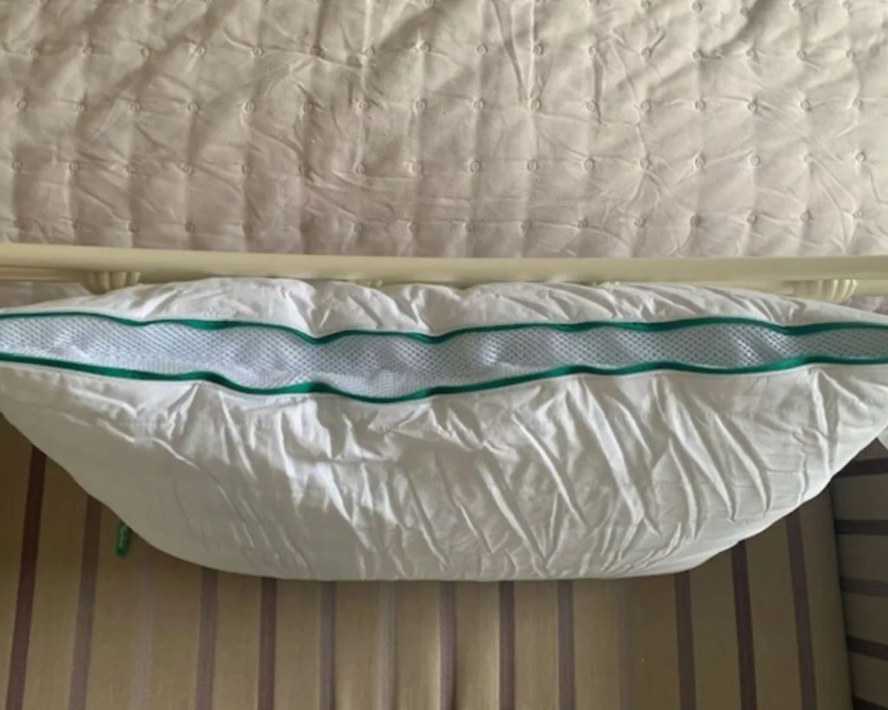A Brooklinen Marlow Pillow photographed from above leaning against a white metal endboard of a bed, on a striped ottoman