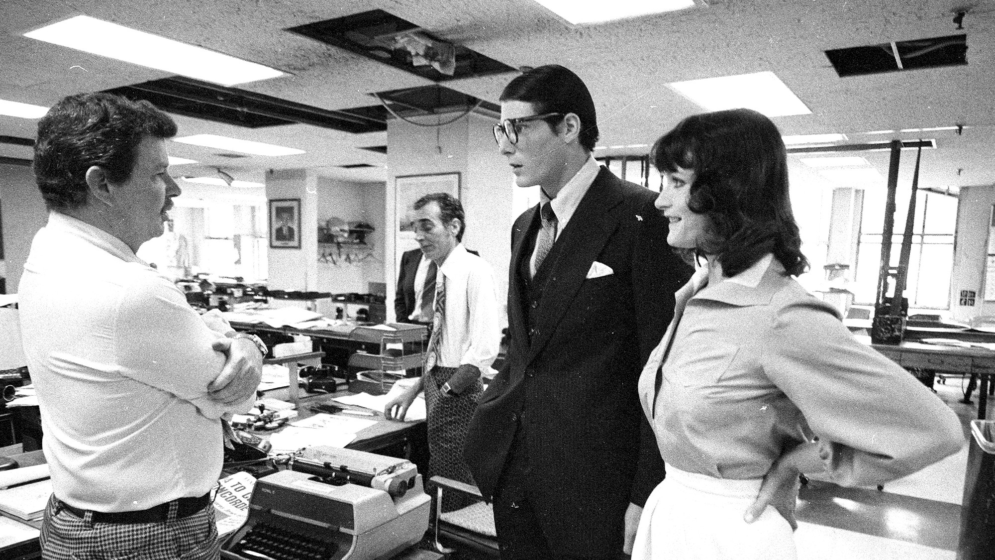 production still in black and white of Christopher Reeve and Margot Kidder during the filming of the first Superman movie. he is dressed as Clark Kent. they are in the newsroom. 