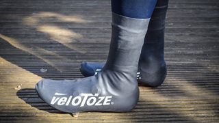 A pair of feet on a wooden deck wearing black veloToze overshoes with white logos