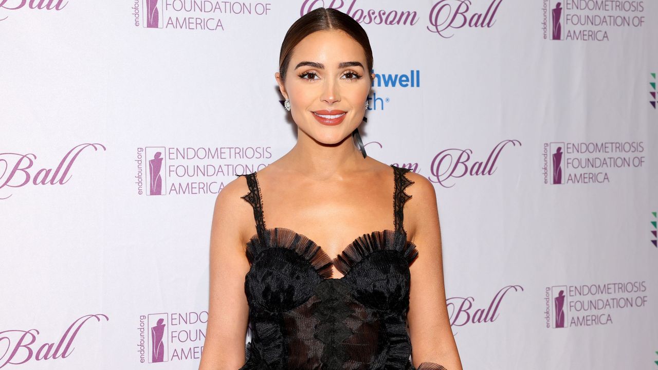 Olivia Culpo, a brunette woman wearing a black lace dress, standing in front of a white background with purple lettering on it