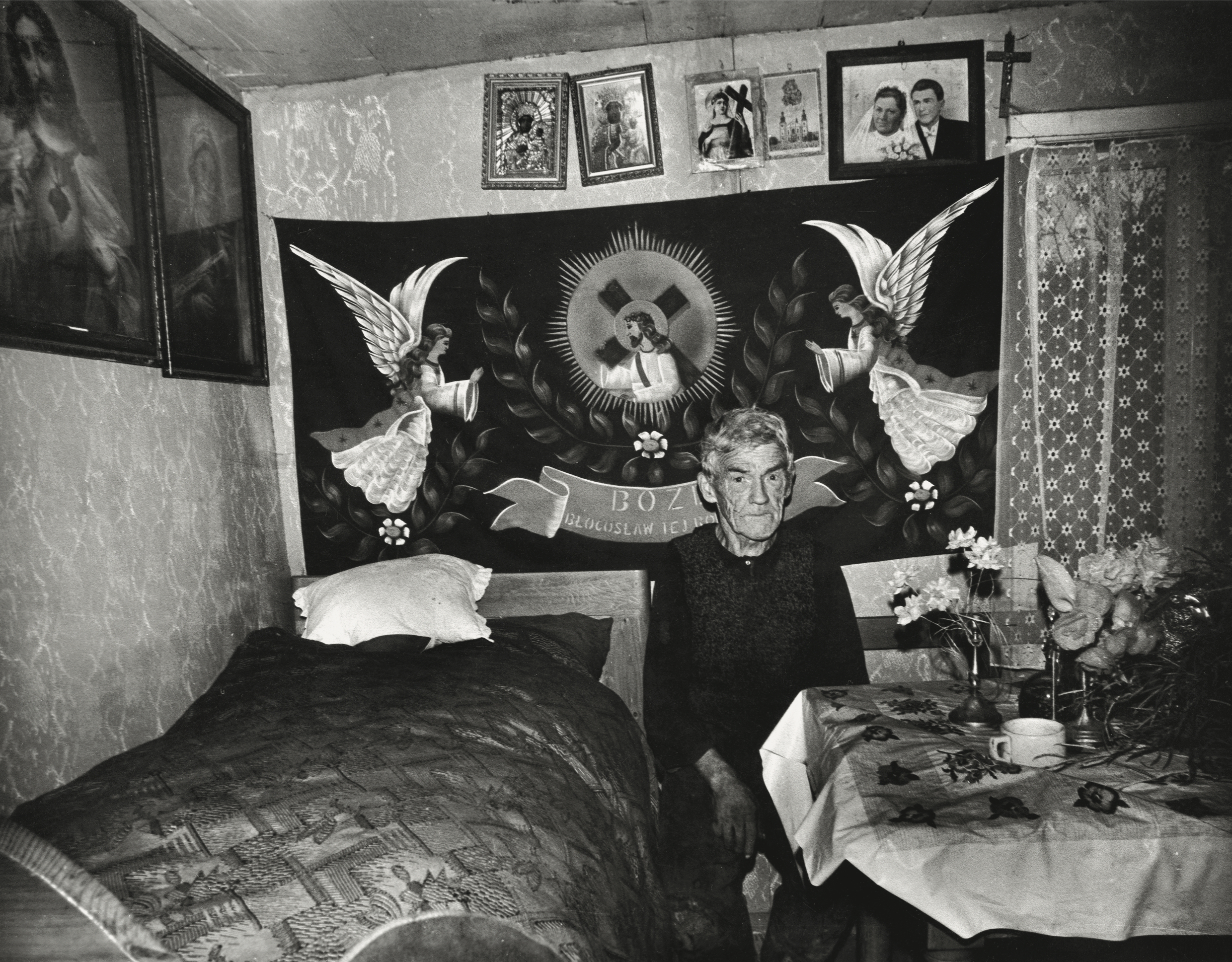 Black and white photograph of an olderman sat in the middle of a bedroom. Family photographsand religious ephemera can be seen on the walls around him