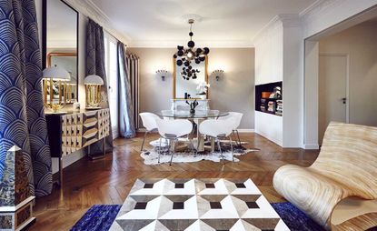 Interior view of the Seine side apartment featuring white and light grey walls, wood flooring, a black chandelier, blue patterned curtains, a round dining table with white chairs, a wooden chair with a curved base, a sideboard with two white and gold lamps on top, mirrors, a recessed storage area with books and rugs