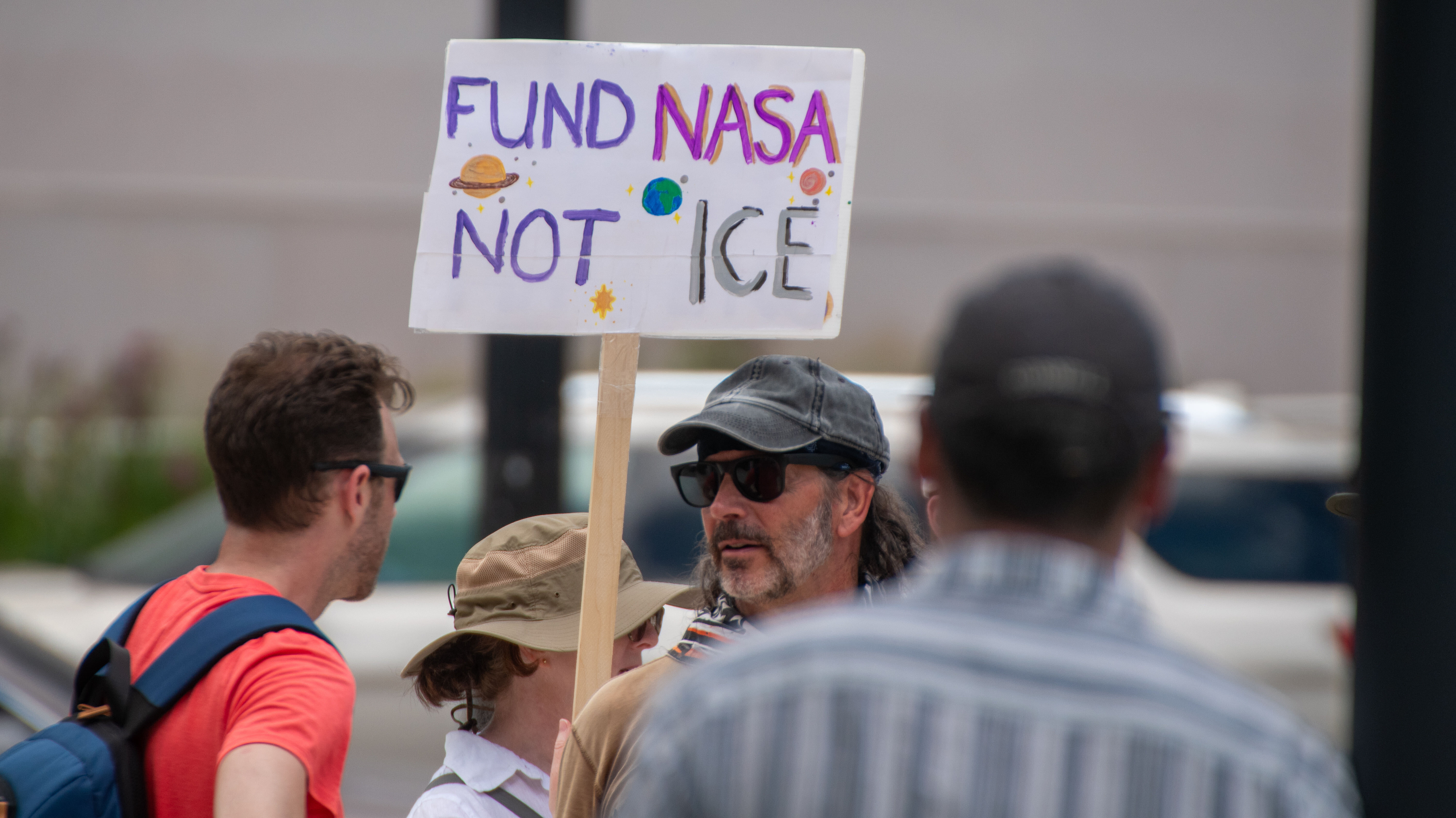 People hold signs outside on a sunny day in protest of NASA budget cuts, July 20, 2025.