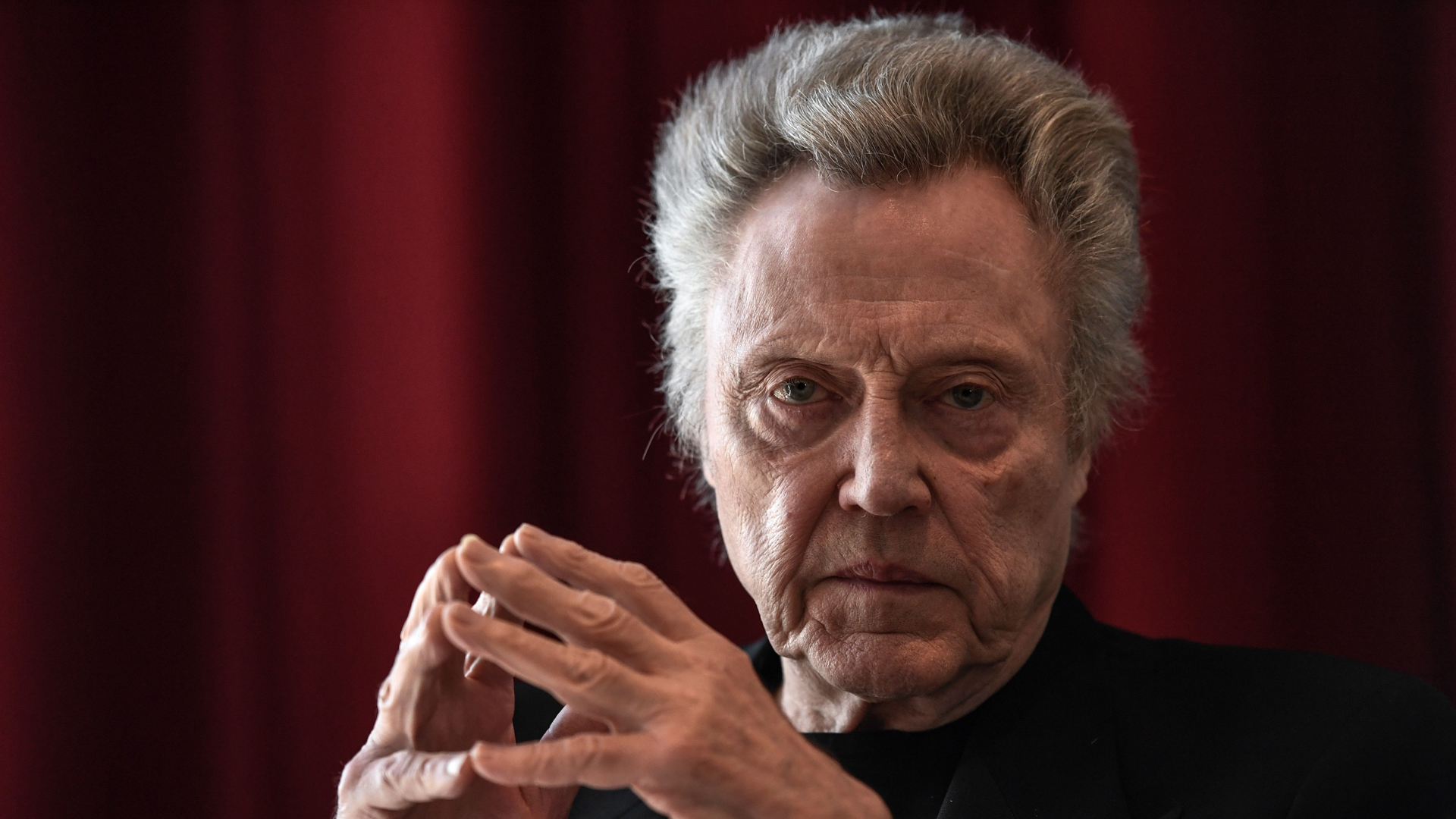 An official image of Christopher Walken staring at the camera with his fingers pressed against one another
