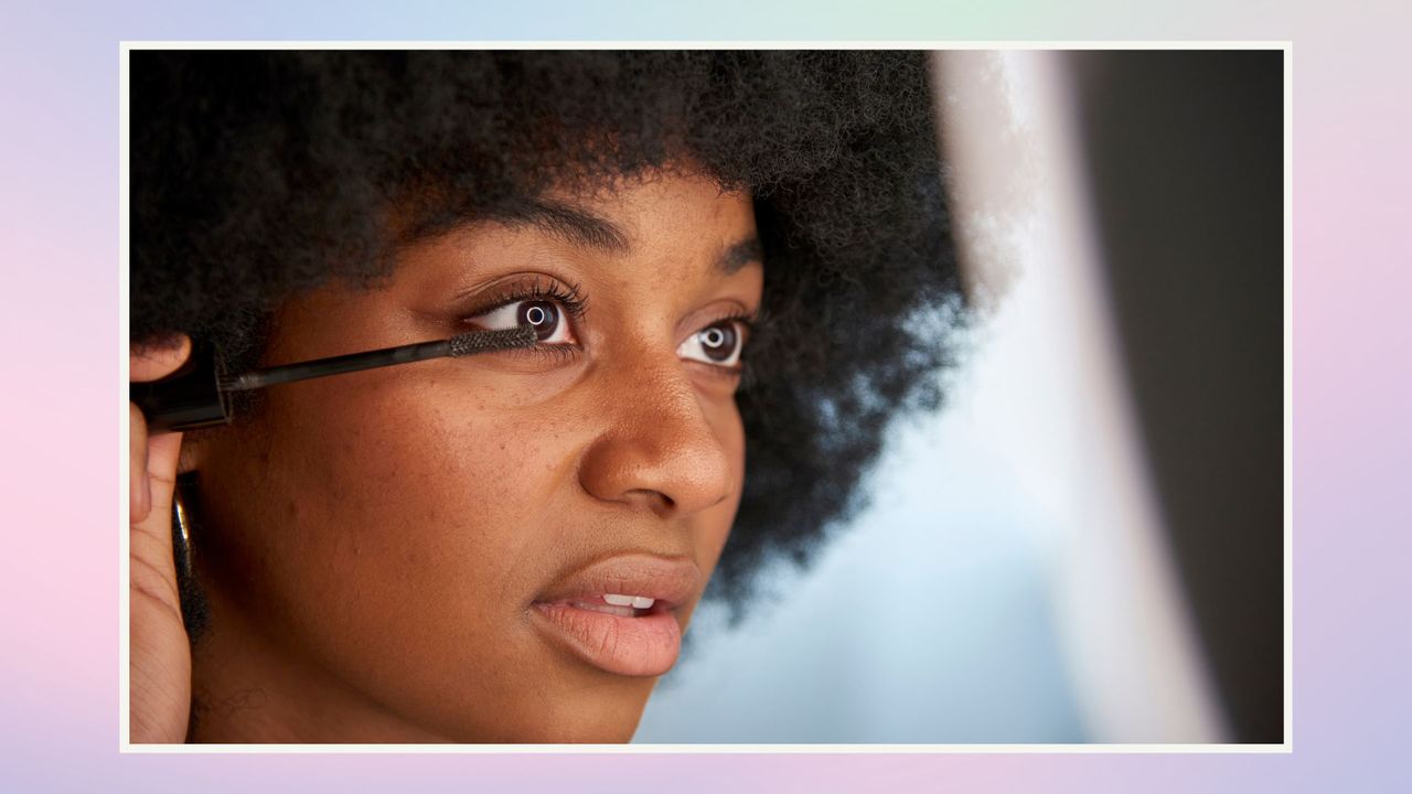 Woman applying mascara to her left eye while looking in a mirror / on a blue, pink and green background