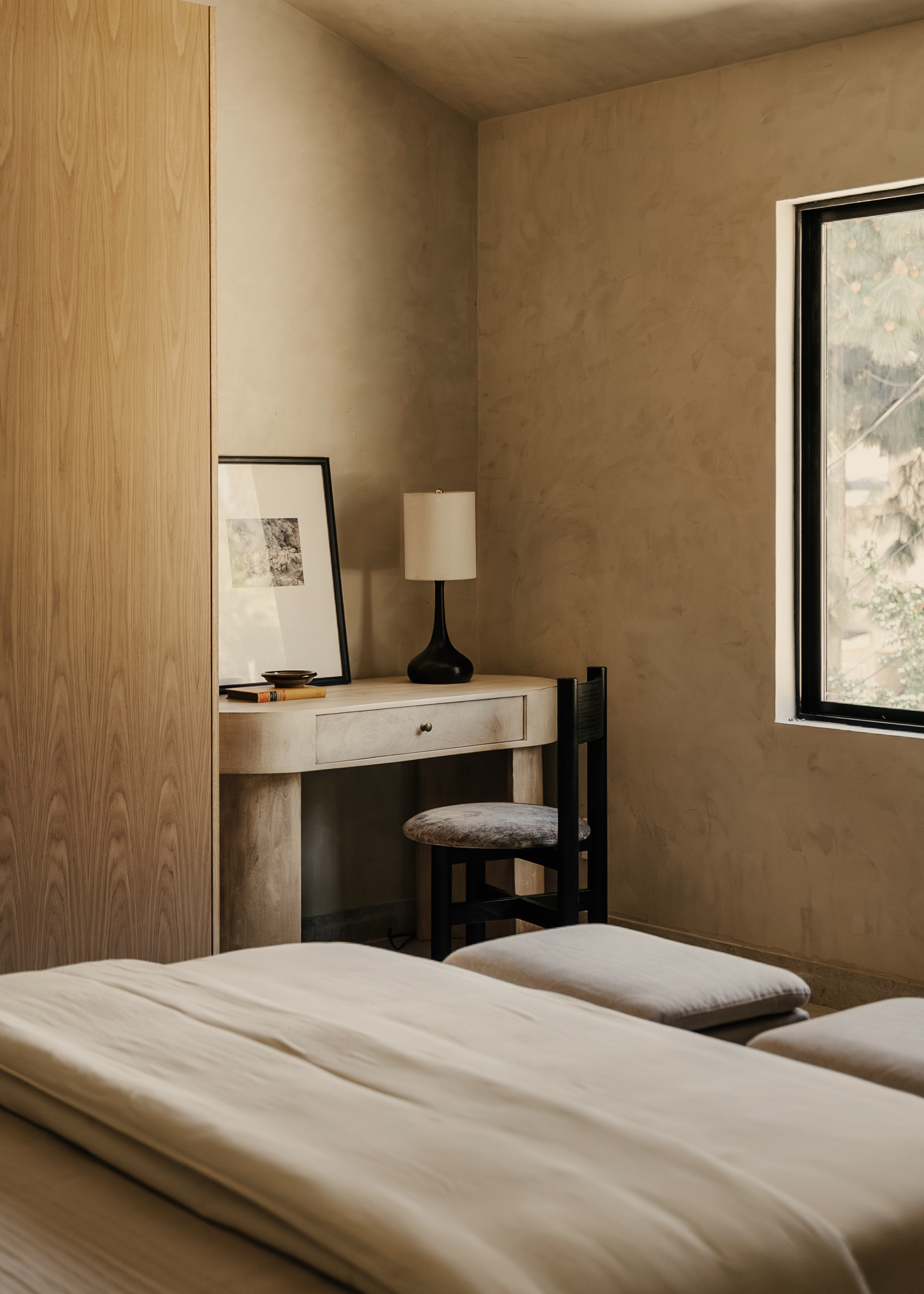 A minimalist bedroom with a desk and a chair by a window. Also pictured are a pair of stools at the foot of the bed.