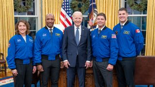 four people in blue flight jackets pose for a picture with a man in a suit in front of an american flag in an ornate office 