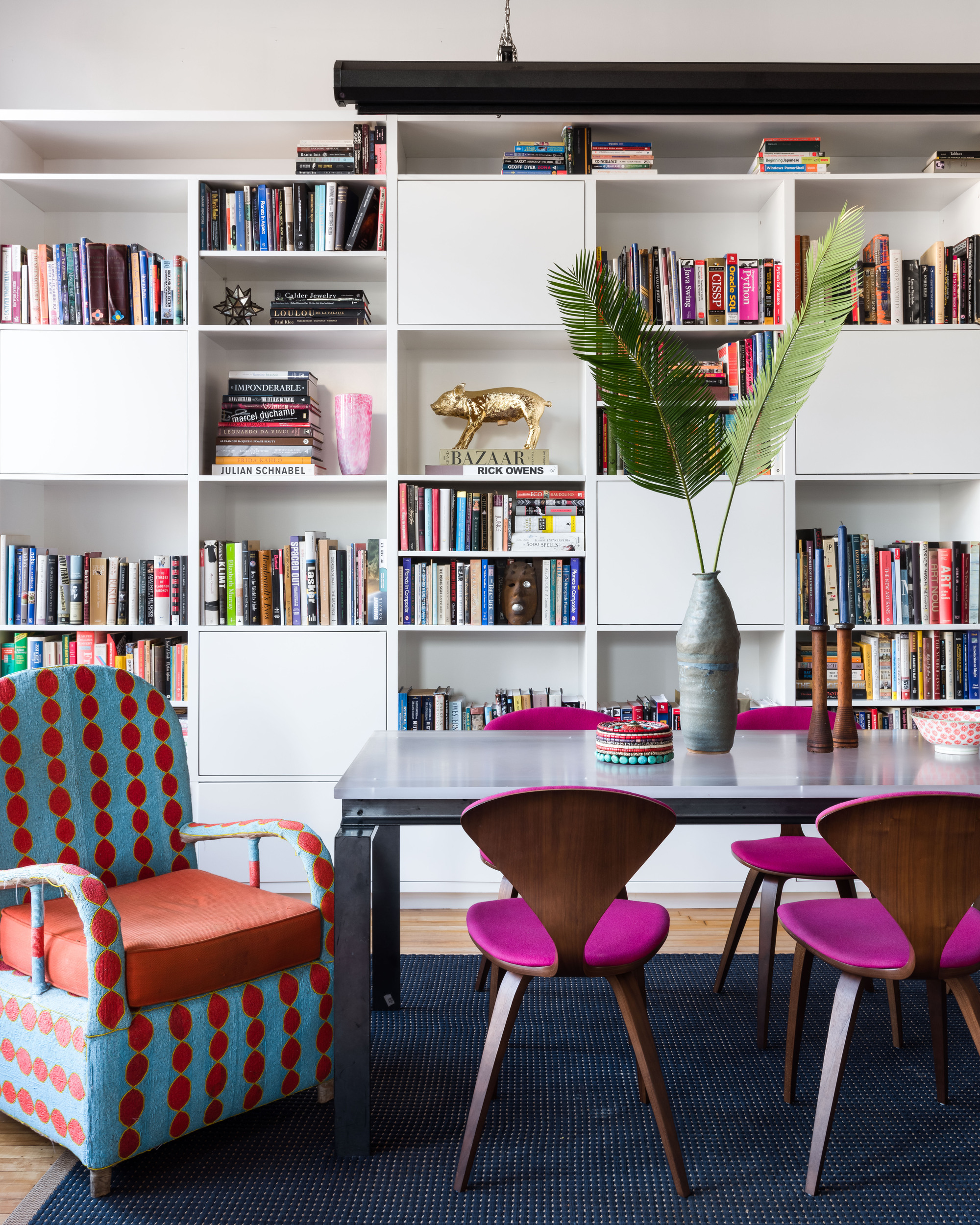 Image of a modern dining room. The back wall is a white floor-to-ceiling bookshelf filled with books. The dining table is on top of a navy rug. There are four magenta and wood dining chairs around the table and a red and blue beaded chair at the head of the table.