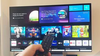 En hand håller upp en fjärrkontroller mot en LG C2 TV som står på en TV-bänk och visar en meny med filmer och serier.