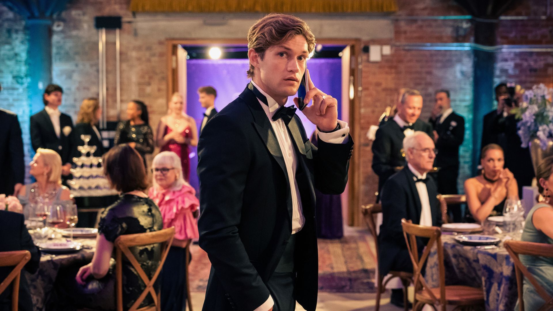 James holds a phone to his ear in the middle of a seated party crowd