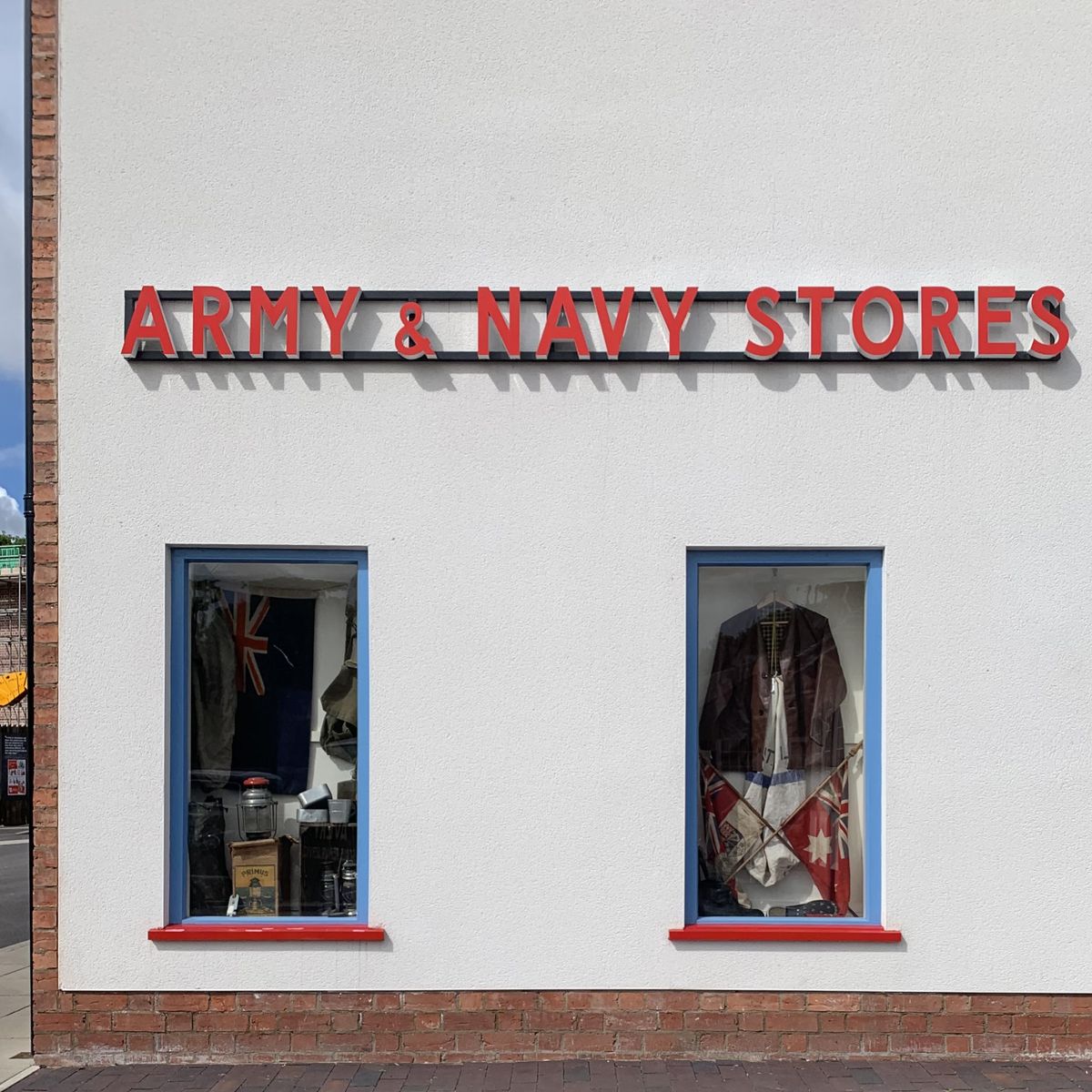 A white shopfront with red "Army &amp;amp; Navy Stores" lettering above two windows displaying vintage military flags and equipment.