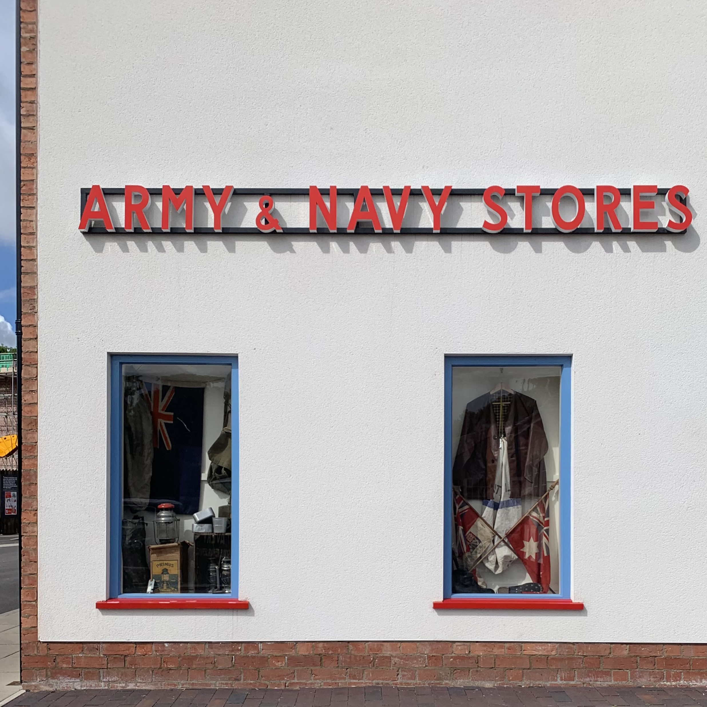 A white shopfront with red "Army &amp;amp; Navy Stores" lettering above two windows displaying vintage military flags and equipment.