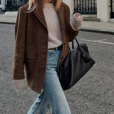 Danish style influencer Jeannette Madsen walking in London wearing a fall 2025 outfit with a brown suede jacket, a neutral crewneck sweater, black leather bag, and straight-leg jeans