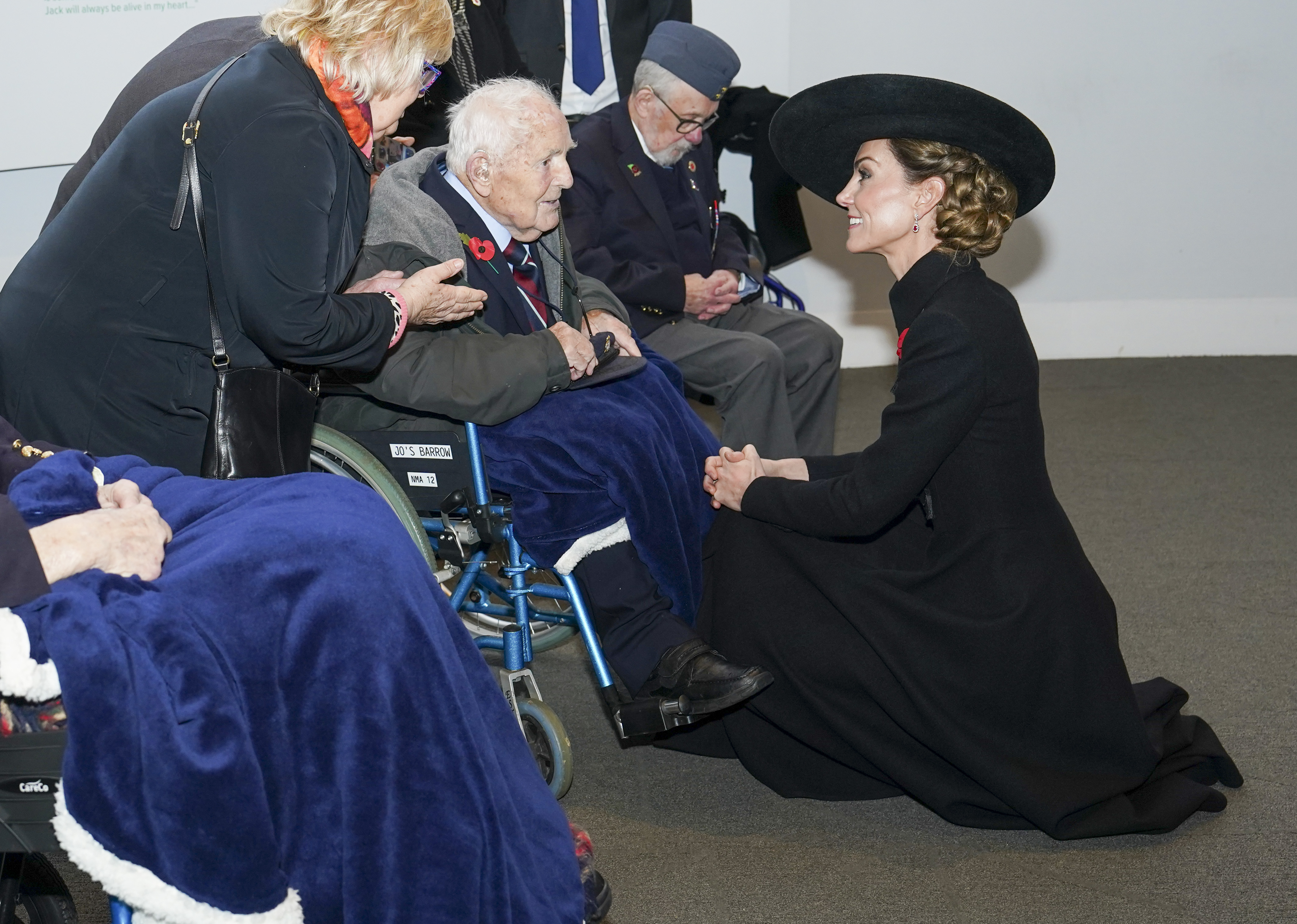 Kate Middleton kneeling down in a black coat smiling at a veteran