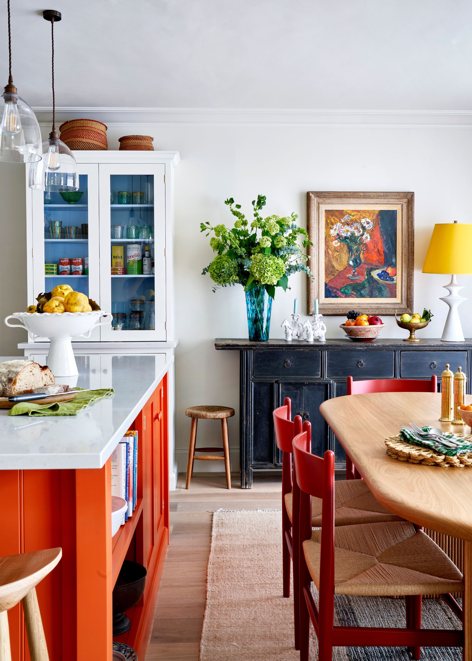 A modern open-plan kitchen with white walls, a wooden dining table with red chairs, and a bright orange kitchen island with white countertops.