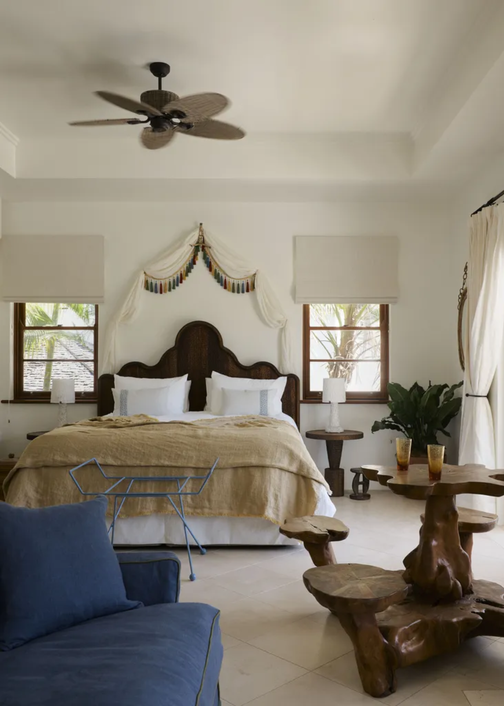 A calming bedroom with overhead draping, a curved headboard, a wooden table with attached seating, a blue sofa and a blue luggage rack