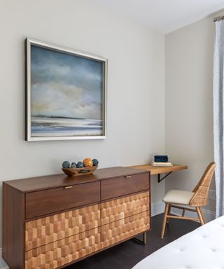 A wooden set of drawers with an oak desk and chair next to it, with a blue wall art print above it