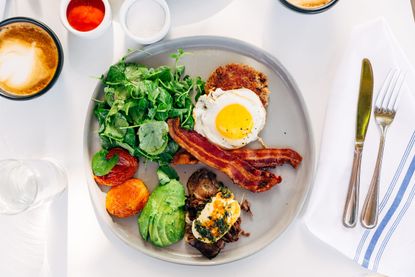 A lazy keto diet breakfast of eggs, avocado, tomatoes, bacon, spinach and mushroom on a plate next to a knife and fork and a coffee.