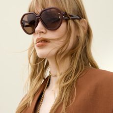 Woman wears oversized brown sunglasses, brown blazer and gold necklace