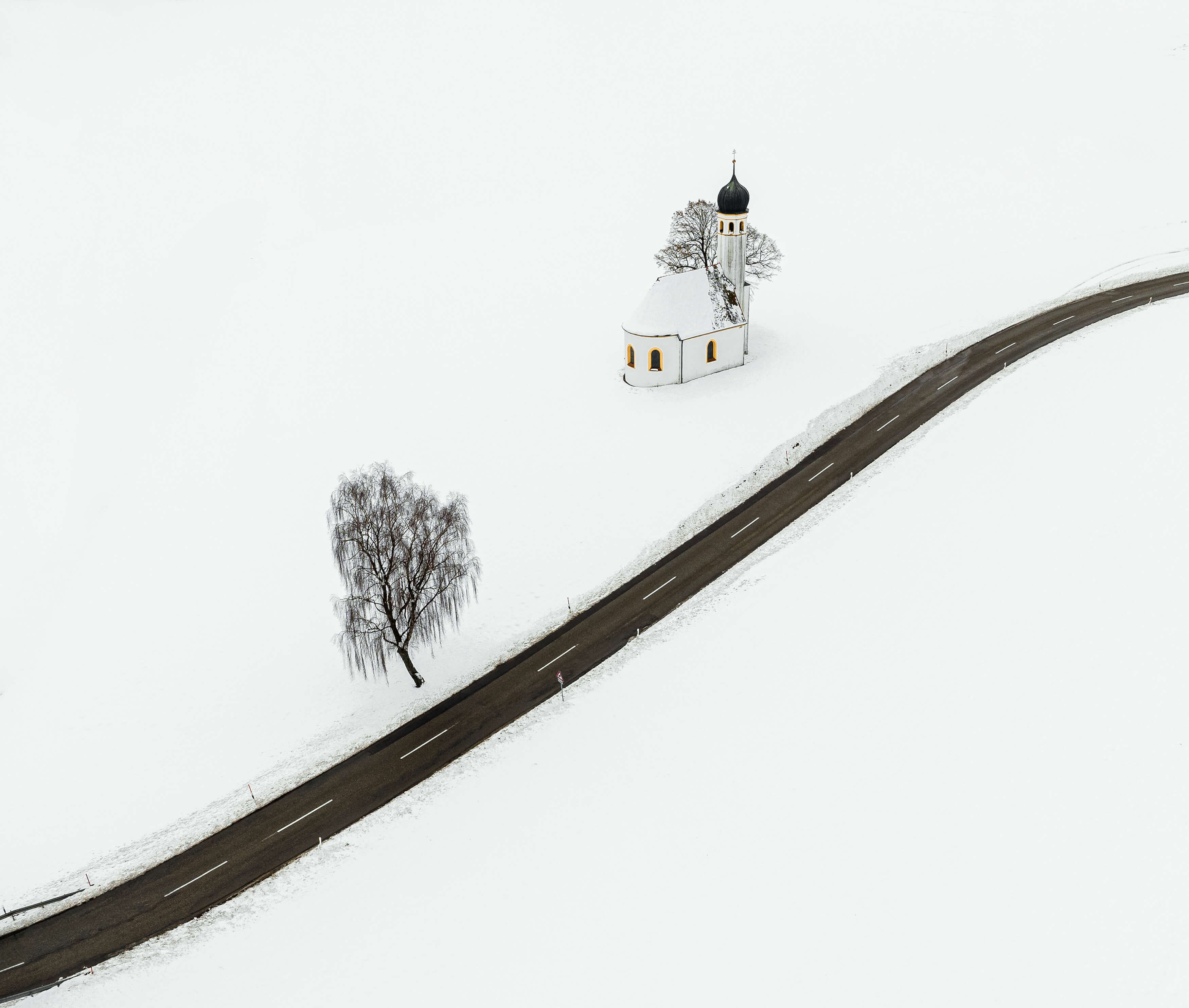 Aerial view of road running through snow-covered field. There is a church and a tree on the left-hand side of the road.