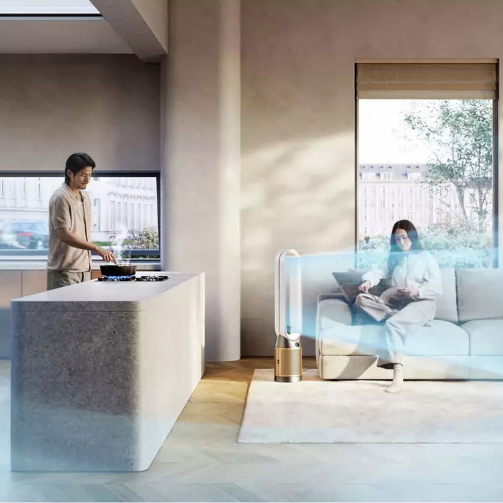 Dyson Find+Follow purifying fan sitting in an open-plan kitchen and living room, with a woman sitting on a grey sofa next to it, and a man cooking in the kitchen