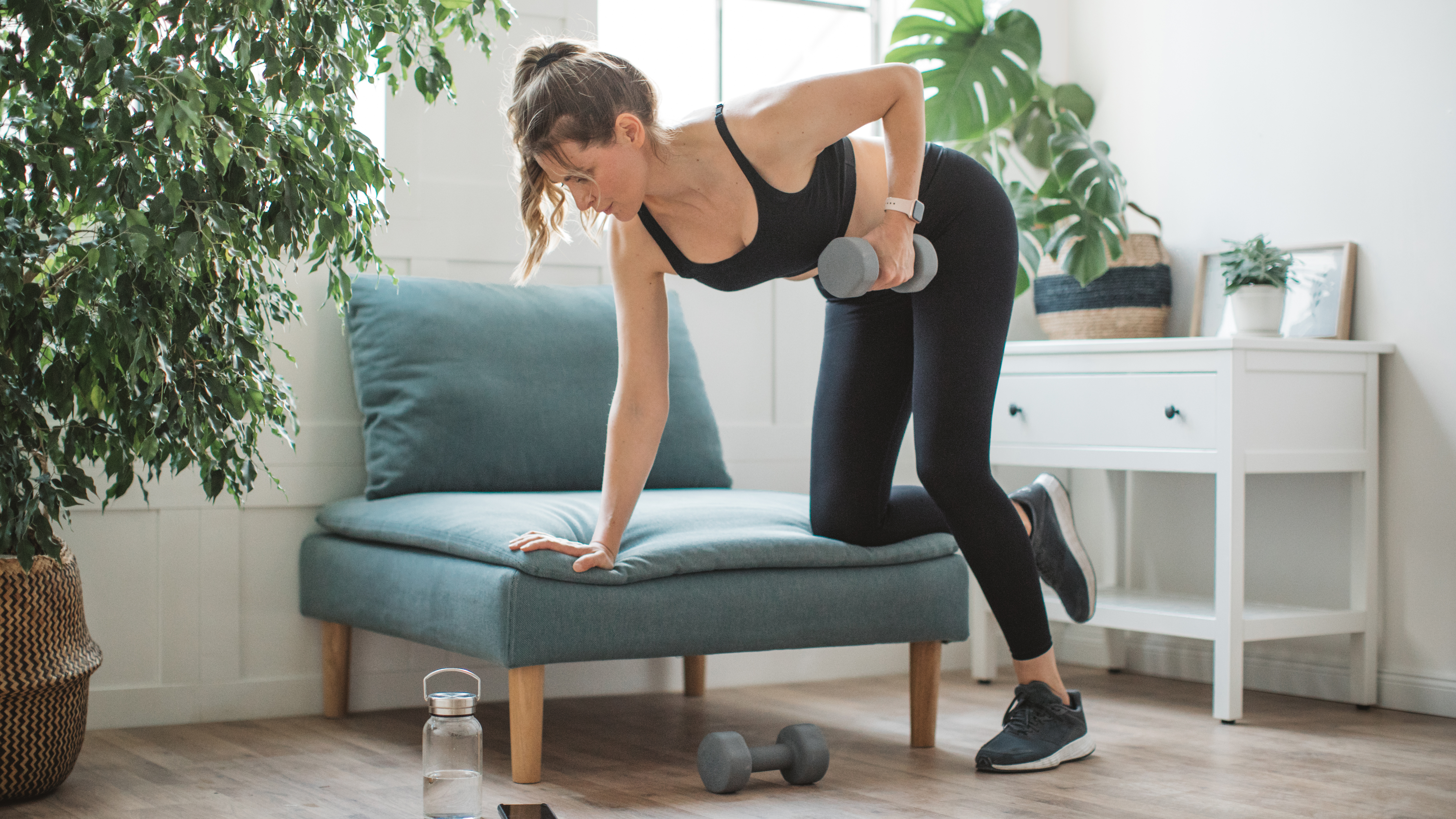 A woman performs a bent-over row with a dumbbell at home on a wide chair. Her right arm and right knee are on the chair, her left leg on the floor, and her torso is bent forward so her back is flat. In her left hand she holds a dumbbell, which she is rowing up to her ribcage. There's a chest of drawers and plant behind her. 