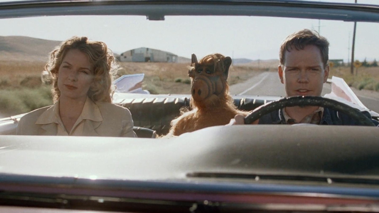 ALF the furry, croissant-nosed alien is sitting in the back seat of a convertible. A male agent is driving and a female agent is in the passenger seat. They are driving along a country highway.