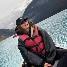 Hailey Bieber wearing a shearling jacket, baseball cap, and sitting in a lake