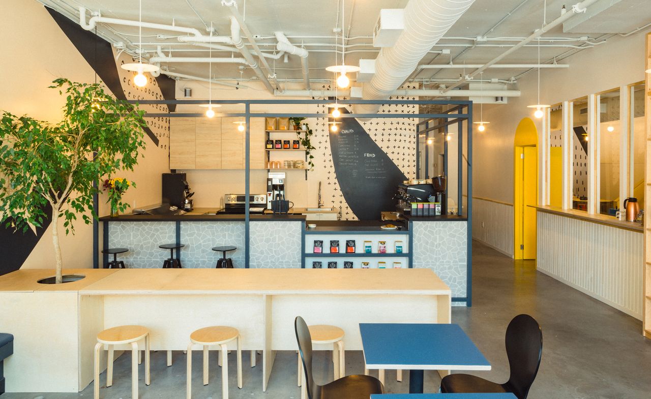 Interior view of the café at Les Fraiseurs featuring light coloured walls, grey floors, pendant lights, a long wooden table with a hole and tree at one end, wooden stools, blue tables and black chairs. There is a partitioned area where food and drinks are served featuring a counter, black bar stools, various appliances, cupboards, shelving and products on display