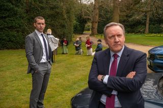 DCI John Barnaby (Neil Dudgeon) sits on the bonnet of his car with his arms folded, DS Jamie Winter (Nick Hendrix) stands slightly behind him with his hands in his pockets. Behind them both is a group of scarecrows arranged on the grass in a semi-circle, kneeling before another scarecrow that has been made to look like a vicar