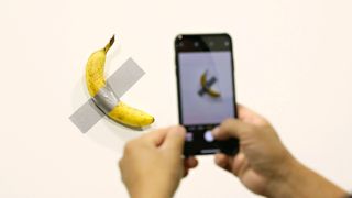 A shot of a banana taped to a wall with a person taking a photo on a phone in the foreground.