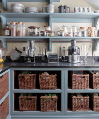 inside shot of pantry designed with blue open cabinets and shelving, black marble countertop, wicker baskets in base units, appliances on worktop and jars and crockery on shelves