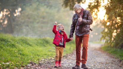A grandfather holds his grandson's hand as they walk down a country path. They are looking at each other and smiling.