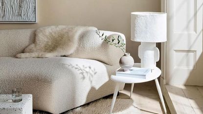 A soft curved cream sofa with a fur throw and a white coffee table with a modern lamp