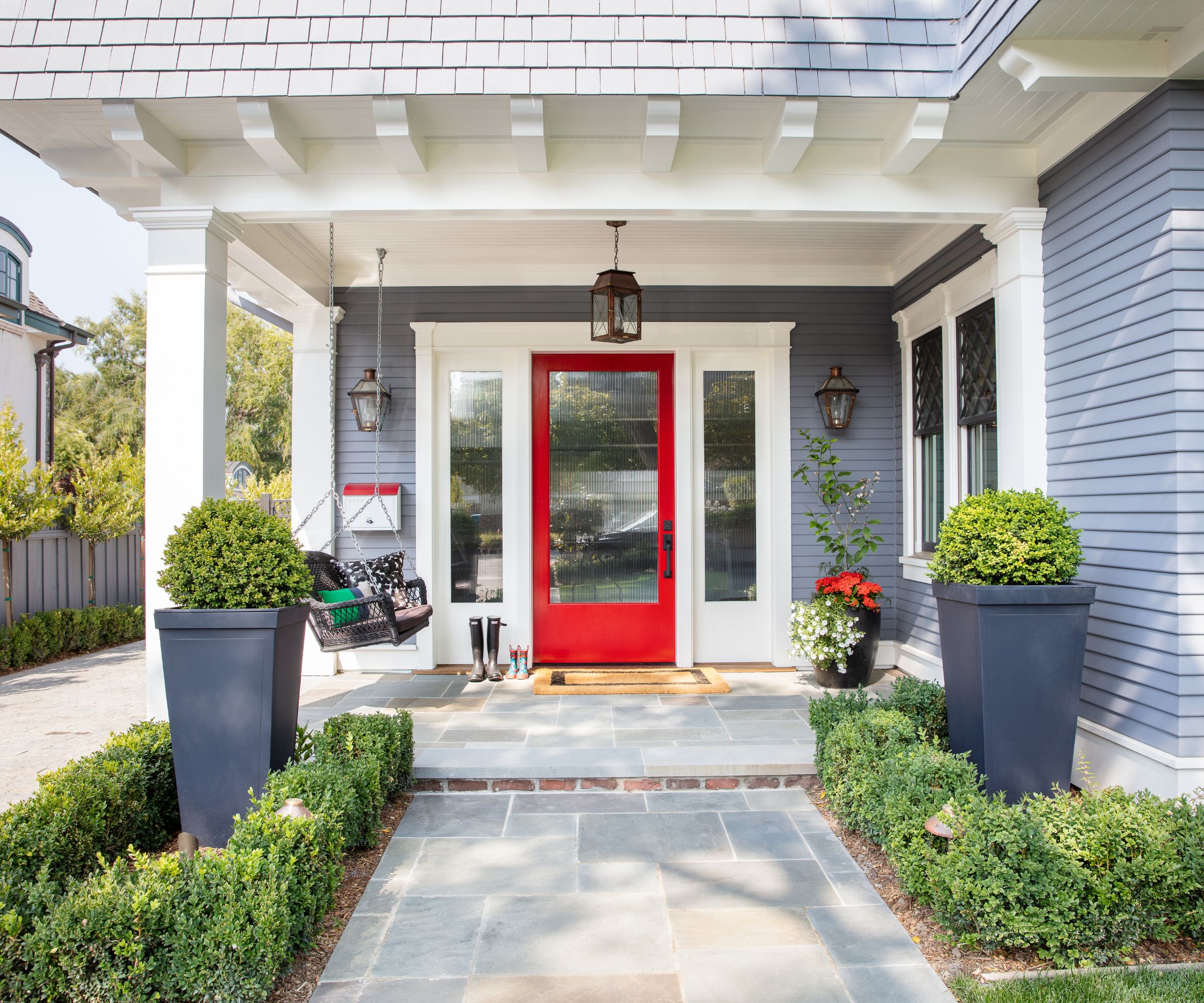 A home exterior painted gray with white trim and a red front door with a glass panel. A porch with lantern-style wall and pendant lights, and potted plants.