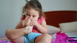 A young girl sitting on a bed smells her own foot.