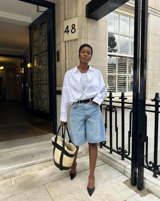 London woman wears long denim shorts, white button down over a tan top, black pointy pumps and a Loewe straw tote bag.