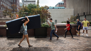 A woman carrying a blue mattress through the street on her shoulder with passers-by watching her