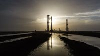 A giant steel rocket stands next to a launch tower silhouetted in front of the sun and reflected in thin pools of water in the foreground.