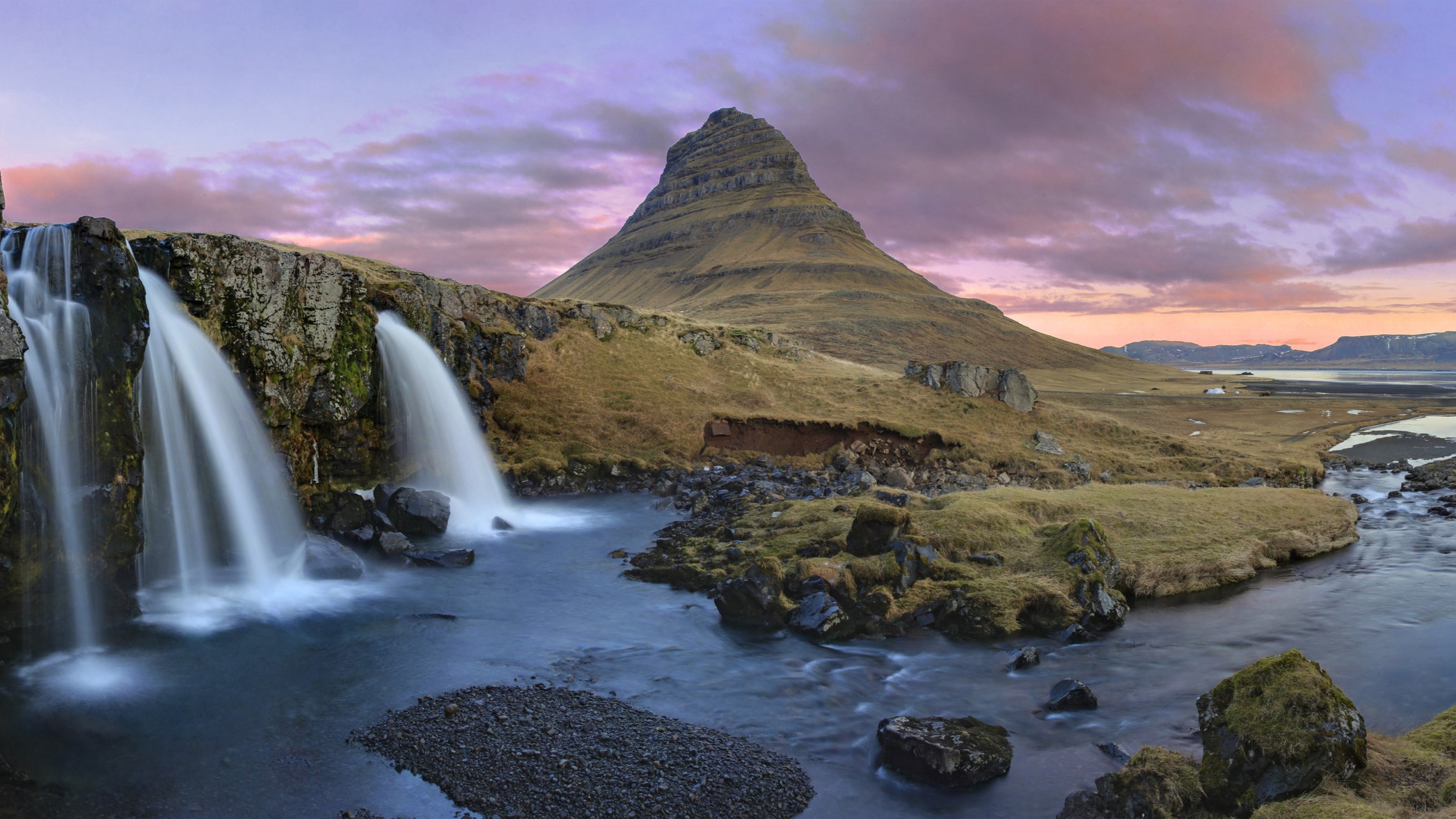 cascading waterfalls in the foreground and a large mountain in the background with a purple pink sky.