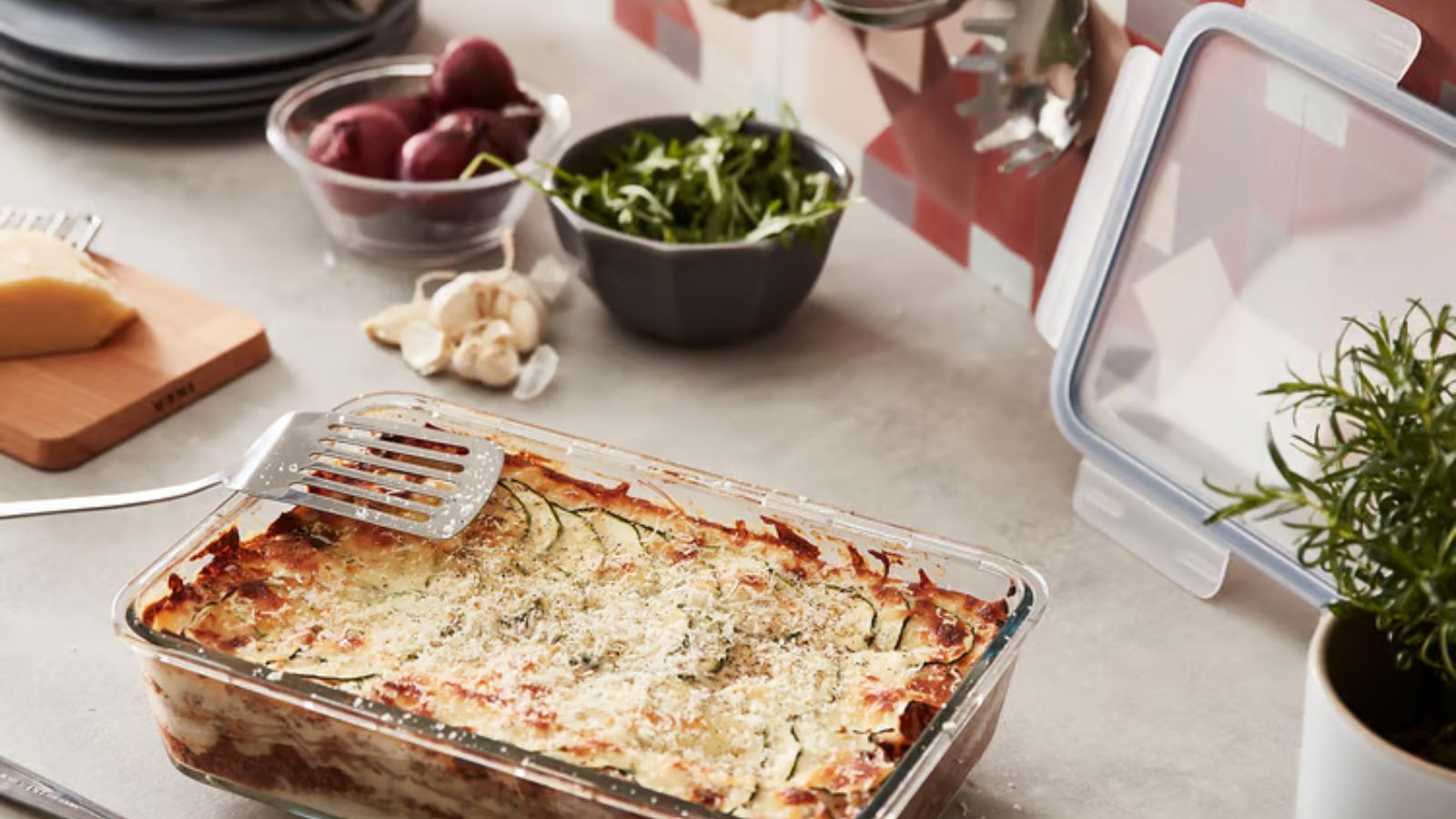 Vegetable lasagne in glass container next to bowls of herbs and spices