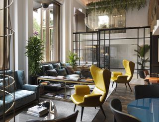 A bright, leafy hotel lobby features colorful 1970s furniture pieces like two futuristic yellow armchairs in leather, along with a series of sleek coffee tables stacked with books and a statement gold minimalist chandelier.