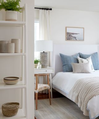 A white bedroom with a blue and white bed, a gray rug, and a shelf with plants and decor on it