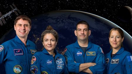 four humans stand in blue jumpsuits in front of a background of a dark earth and shadowed moon, with a small ISS in the top left. 