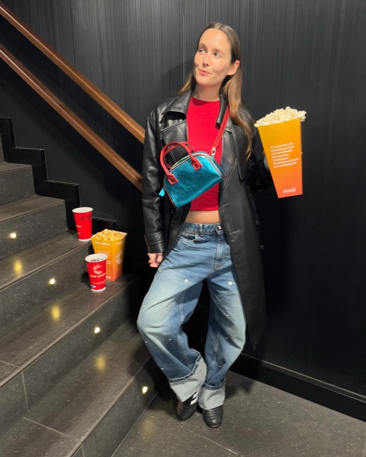 Helena wears a leather long coat, red tee, baggy jeans, Adidas sneakers, and a crossbody bag.