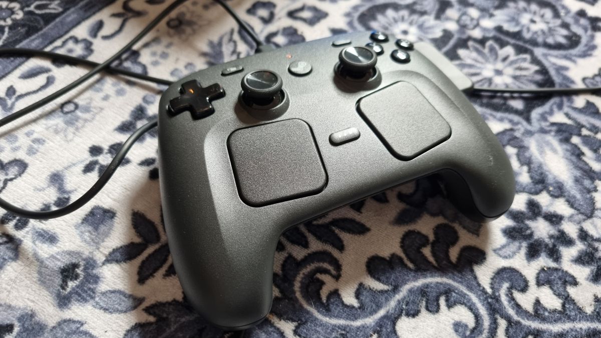 A Steam Controller sitting on a rug, attached by cable to a Steam Machine out of shot