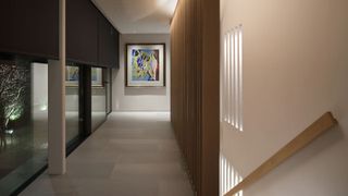 A modern hallway with soft, layered lighting. Recessed ceiling spotlights and vertical wall lights create a warm glow, highlighting a wooden slatted feature wall and a large colourful framed artwork at the end of the corridor
