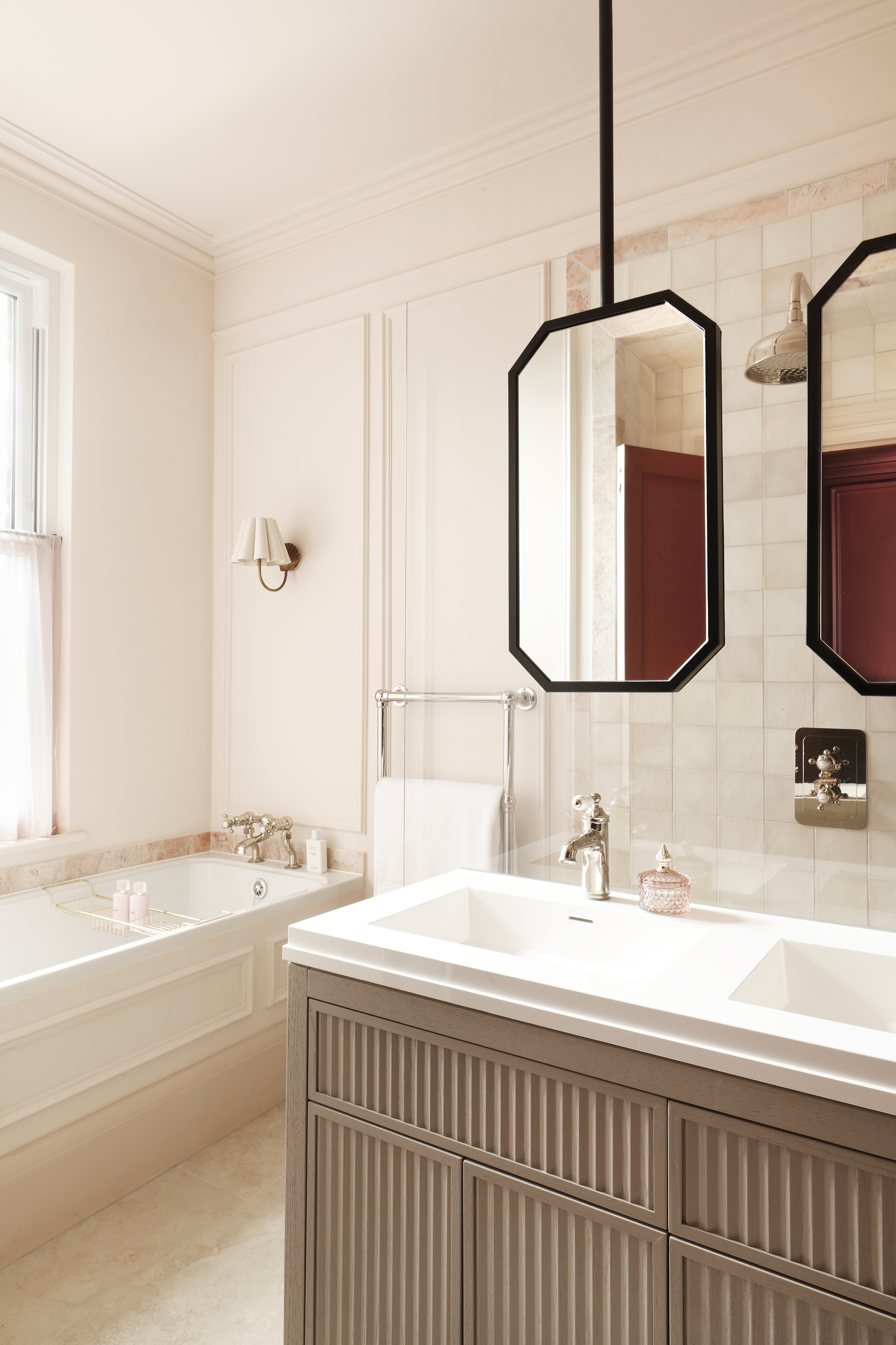 Image of an off-white/light pink bathroom with a tub and an island a double sink unit that has suspended mirrors hanging above it.